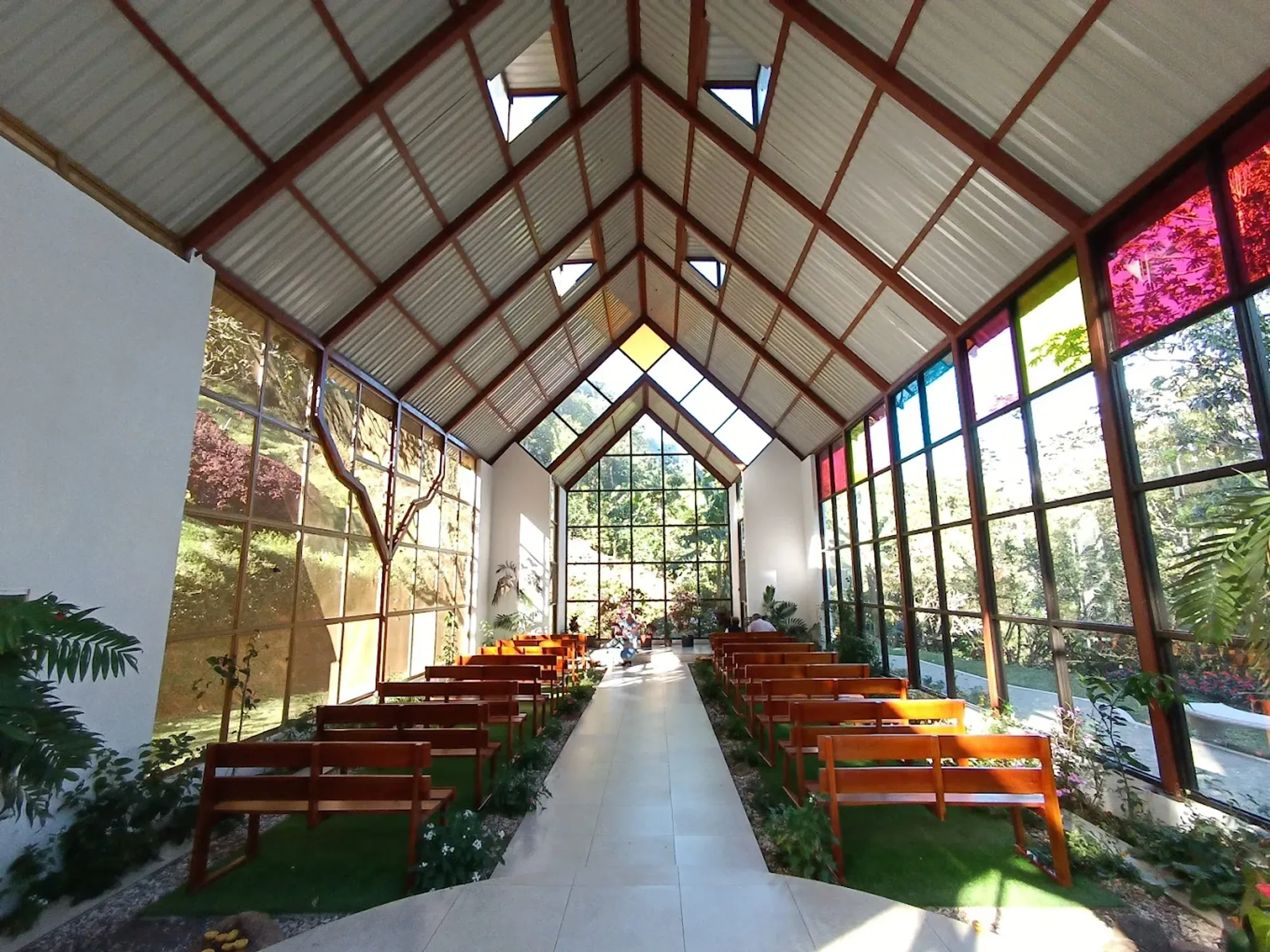 Interior de la capilla: estructura de vidrio y madera con vitrales de colores, bancas de madera y jardín a los costados.